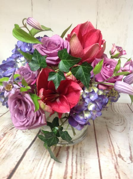 Low glass vase of purple roses, blue hydrangeas, and red amaryllis blooms in a compact arrangement