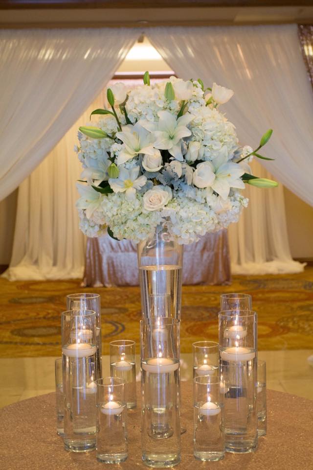 White floral centerpiece in a tall glass vase with candles around it