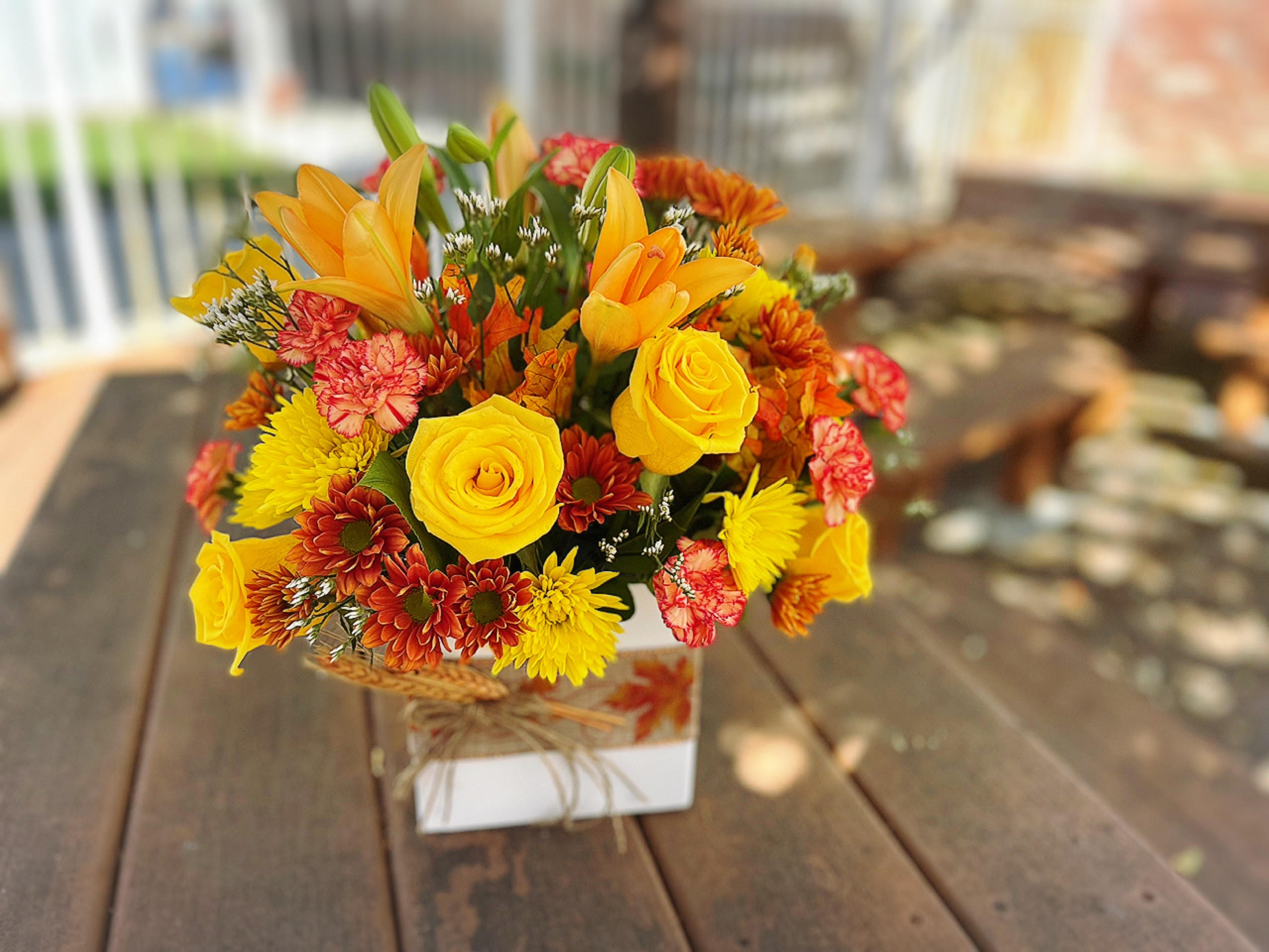 Autumn Present  - - 1 stems Orange Lilies - 6 Yellow Roses - 2 Stems Orange Mini Carnations - 4 Stems Yellow and Orange Daisies - Fall Leaves - Fillers and Greenery - White Decorative Container