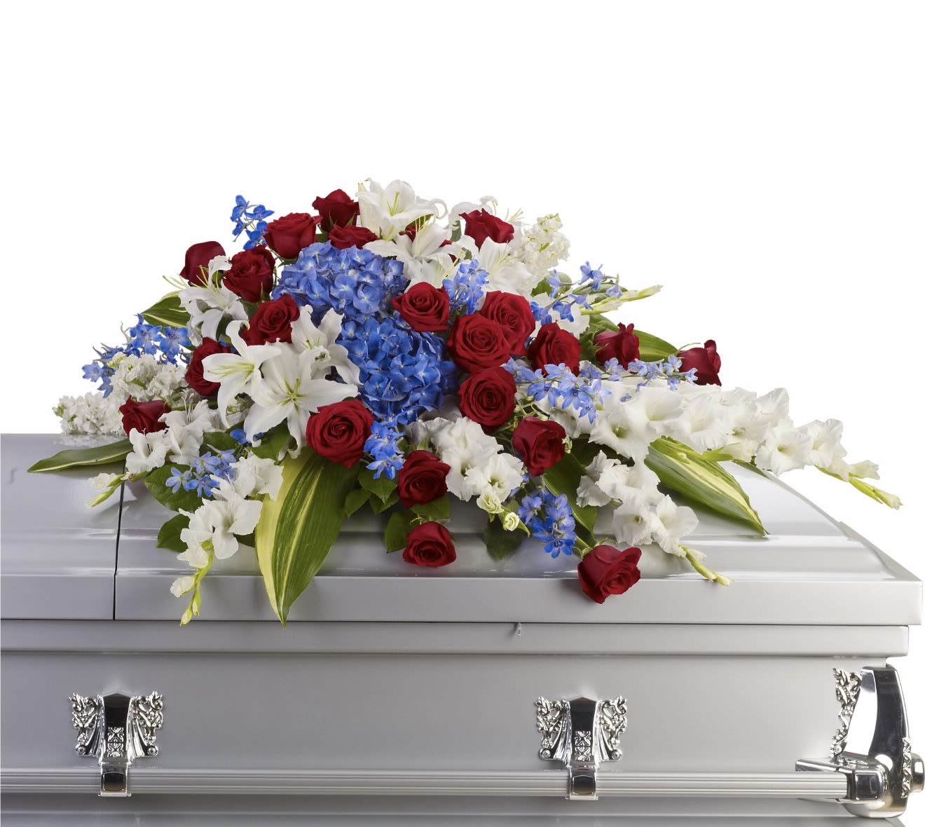 Distinguished Service Casket Spray - A bold tri-color display anchored by dense clusters of red roses, white lilies, and vivid blue hydrangea-like blooms, this large arrangement reads with formal, patriotic grace perfect for a sympathy or funeral setting. The strong red, white, and blue palette conveys honor and respect, while the white lilies add a soft, comforting presence that helps the recipient feel seen and remembered.  Generously layered and wide in shape, blooms are arranged in broad, cascading groupings with some stems and flowers extending outward for a full, flowing silhouette. Lush greenery frames the colors and adds texture throughout; expect a substantial, low-profile design that presents beautifully at a memorial service. This piece is intended to offer a dignified expression of sympathy and tribute for someone who served or for occasions where you want to convey deep respect and heartfelt remembrance.