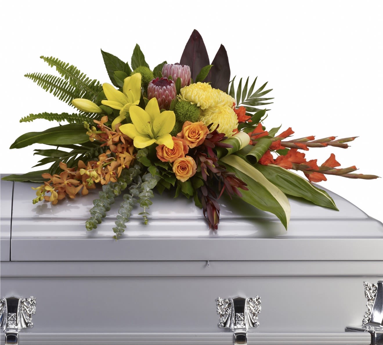 Island Memories Casket Spray - Vivid yellow lilies and bright orange roses meet coral-orange orchid sprays and red tropical accents in a broad, asymmetrical casket spray designed as a heartfelt funeral or memorial tribute. This flower arrangement for a casket or memorial service delivers a warm, uplifting impression-perfect for celebrating a life with tropical color and radiant energy that comforts the person you're honoring.  Built low and expansive to rest across a silver casket lid, the composition pairs broad tropical foliage and dark upright leaves for contrast, letting the blooms extend outward in a dramatic, formal presentation. Colors include yellow, orange, red, and lush greens; flowers shown are lilies, roses, orchids and complementary tropical elements arranged to read beautifully from the front and sides of the casket.  For family and friends seeking a meaningful way to express celebration, remembrance, or aloha, this spray conveys warmth and vivid remembrance at the service. Sized to cover the top of a standard casket, it arrives fully assembled and ready for placement by our professional floral team.
