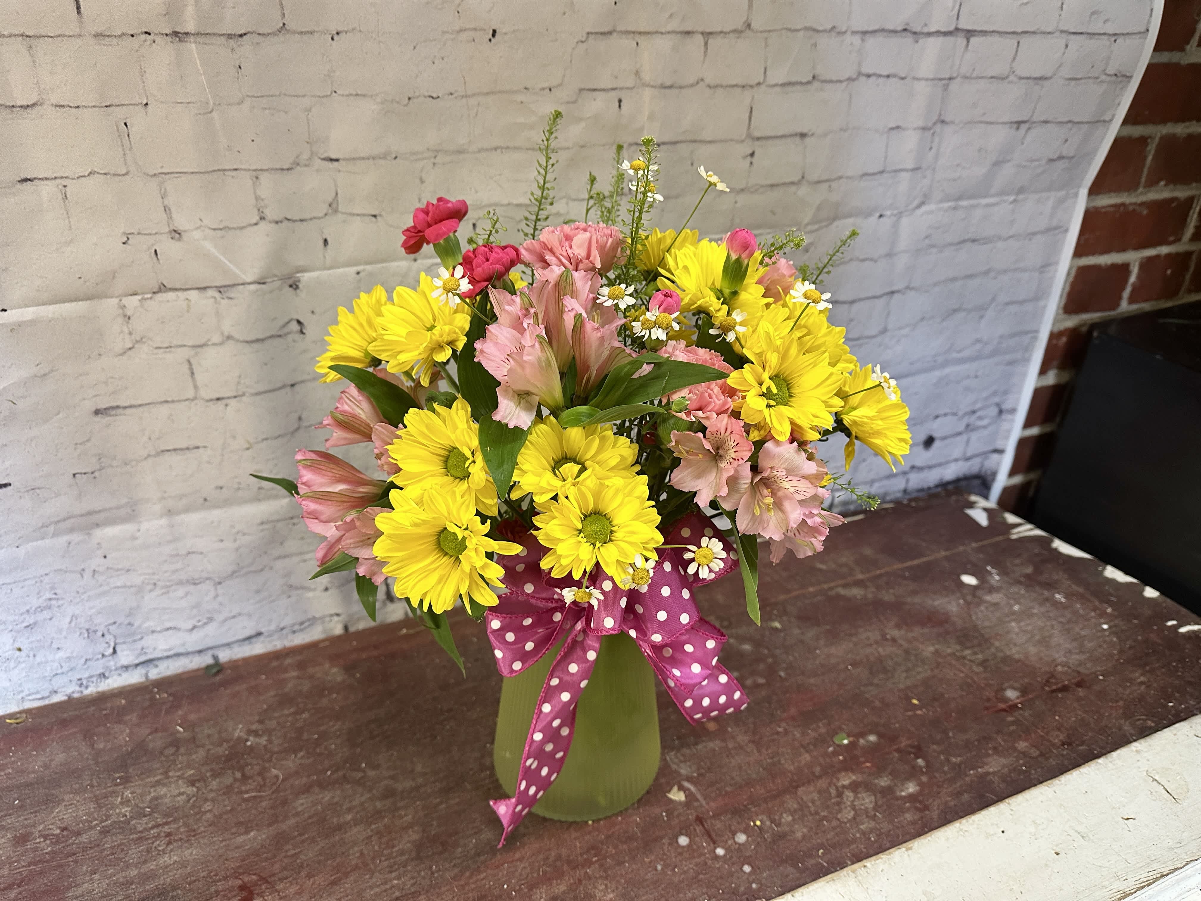 Bouquet of yellow daisies and pink alstroemeria in a green vase with a pink polka-dot ribbon