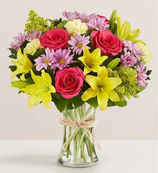 Bouquet of pink roses, yellow lilies, and lavender daisies in a glass vase
