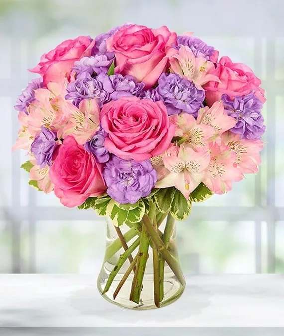 AVA Mother May I - A feminine delight to have as the center to any room Pink roses, lavender carnations, and pink alstroemaeria in a clear gathering vase