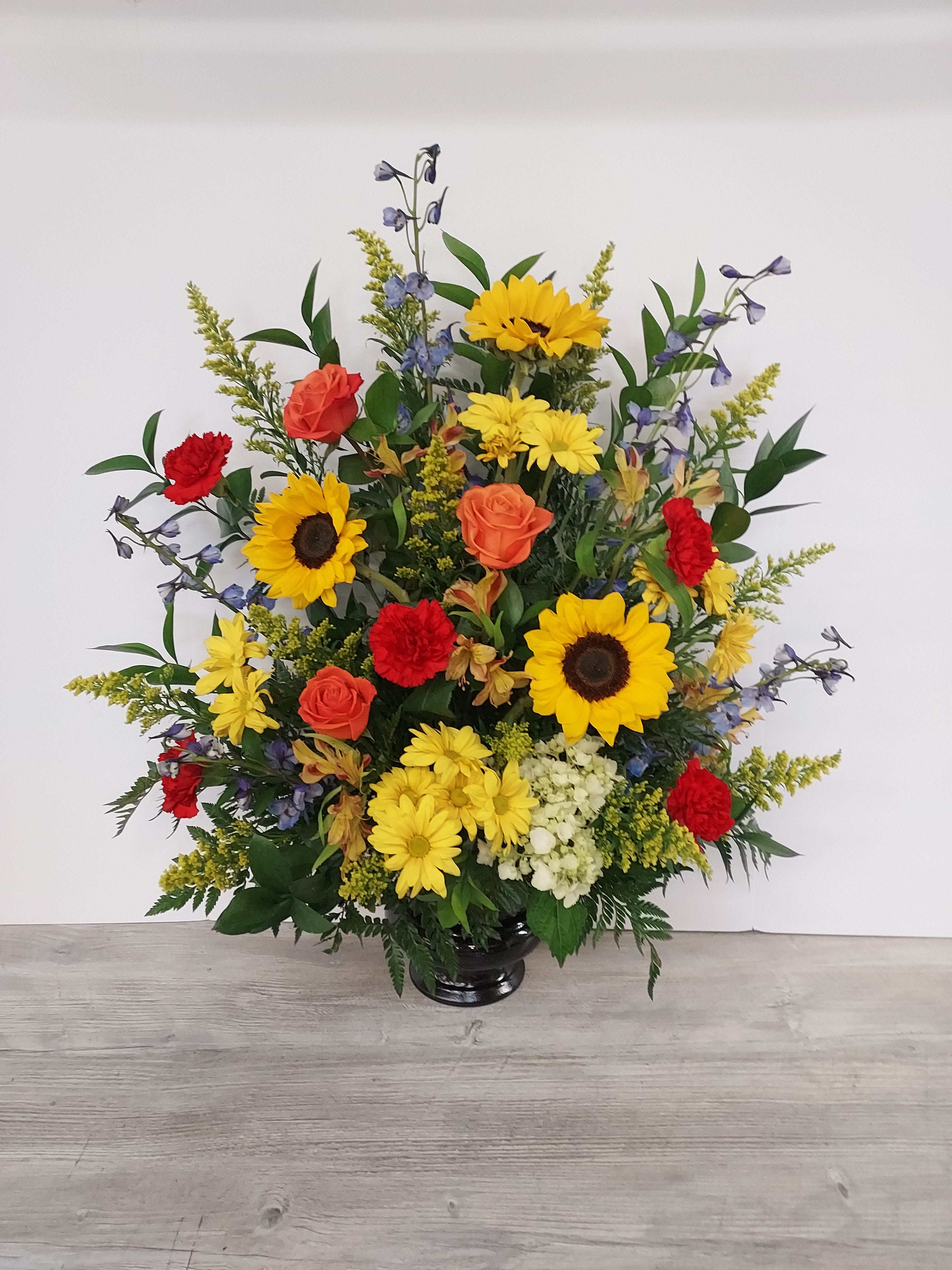 Tall arrangement of sunflowers, yellow daisies, orange roses and red carnations in a dark vase