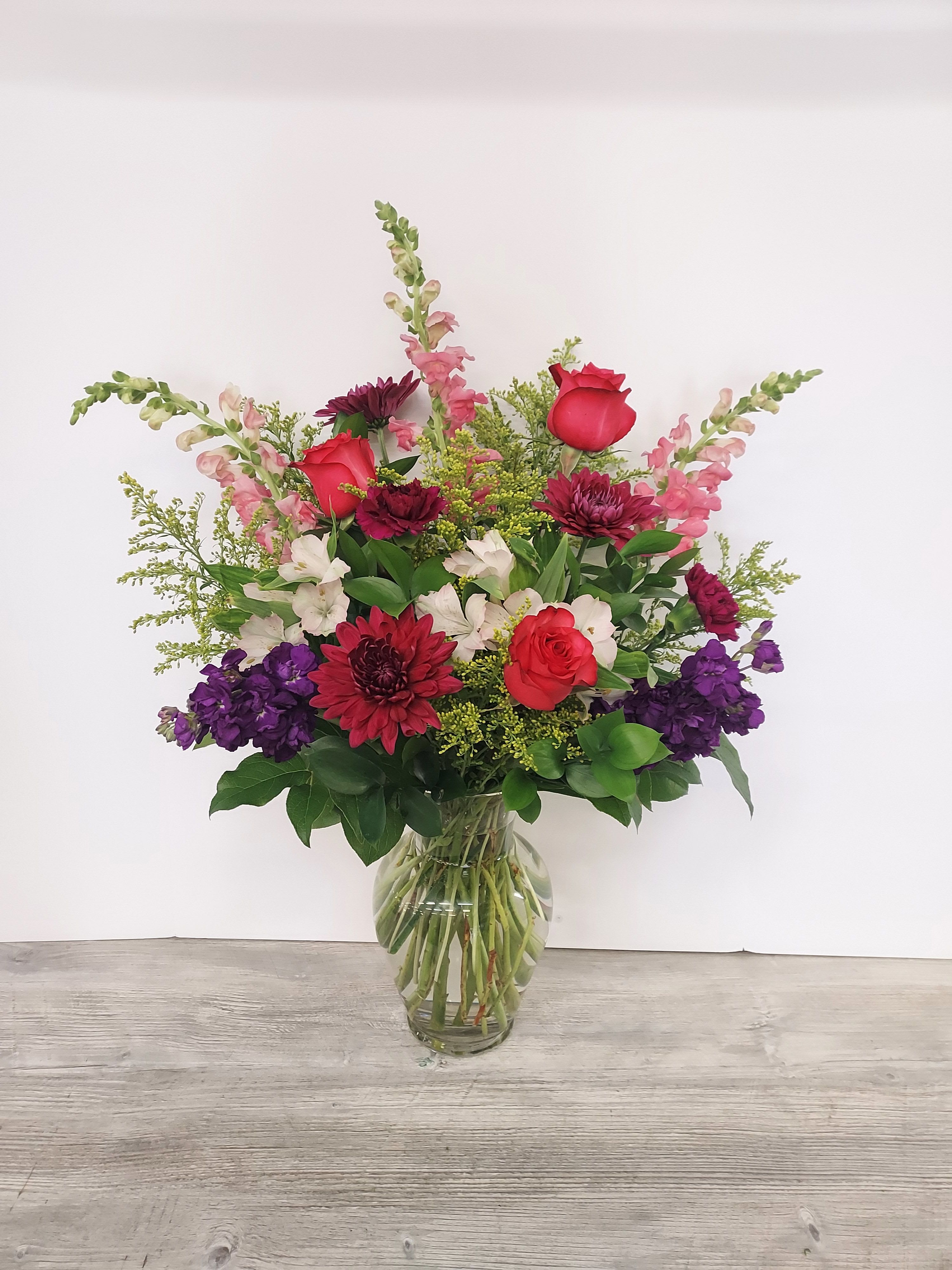Tall mixed arrangement of red roses, burgundy mums, pink snapdragons and purple blooms in a clear glass vase