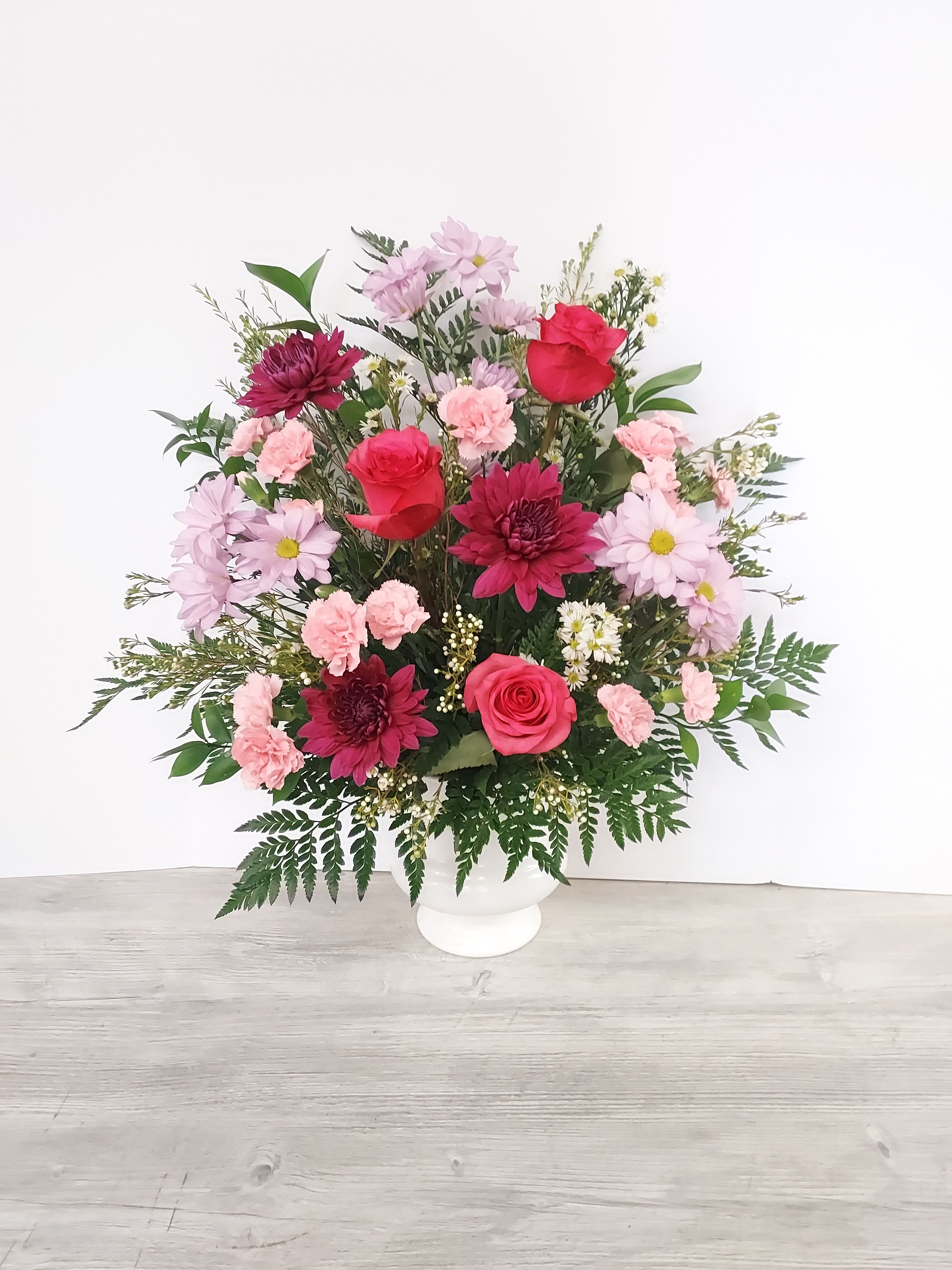 Tall arrangement of red roses, pink carnations, and mixed blooms in a white pedestal vase