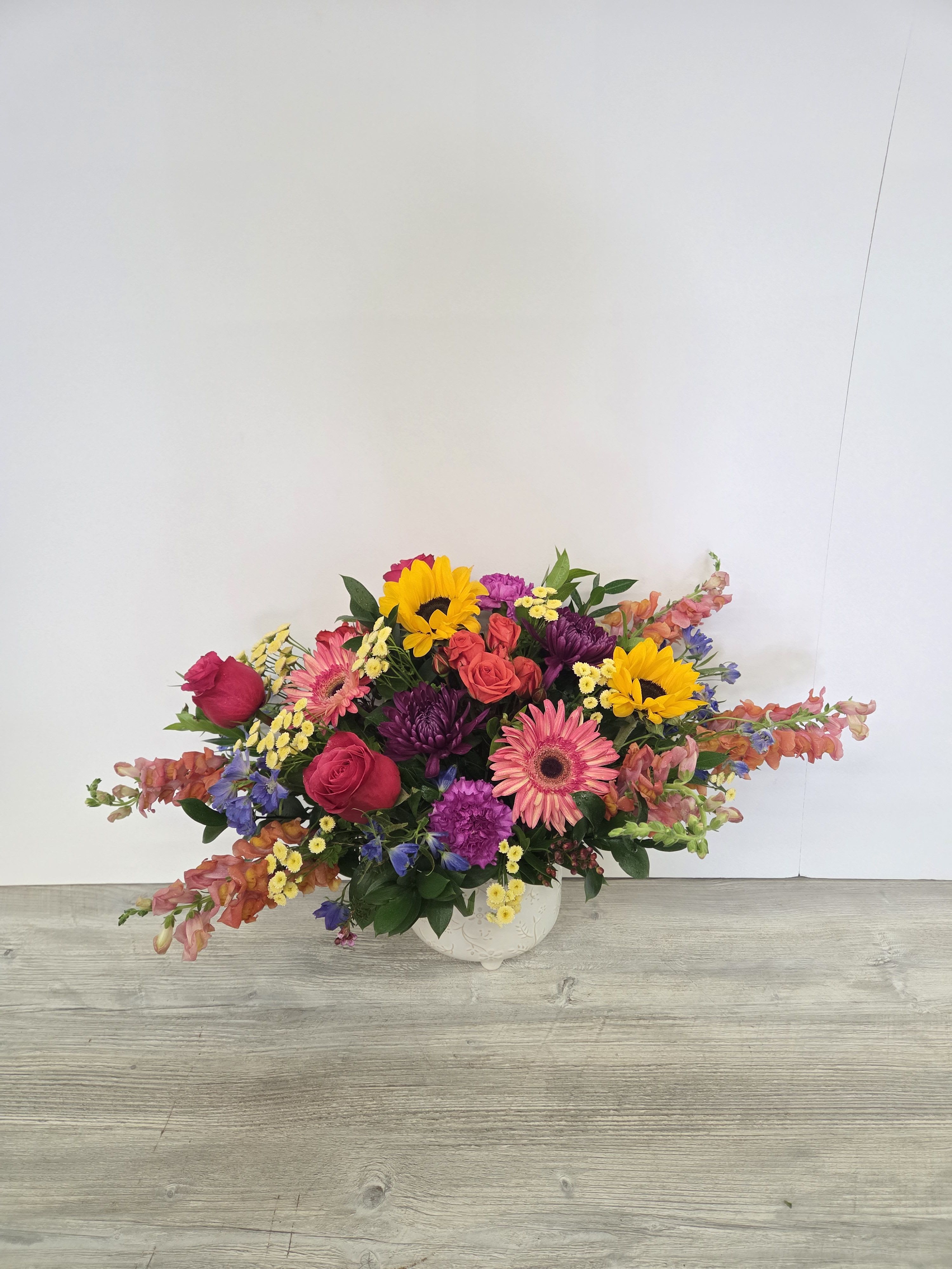 Low colorful arrangement of roses, gerbera daisies, sunflowers, and mixed blooms in a white ceramic bowl.