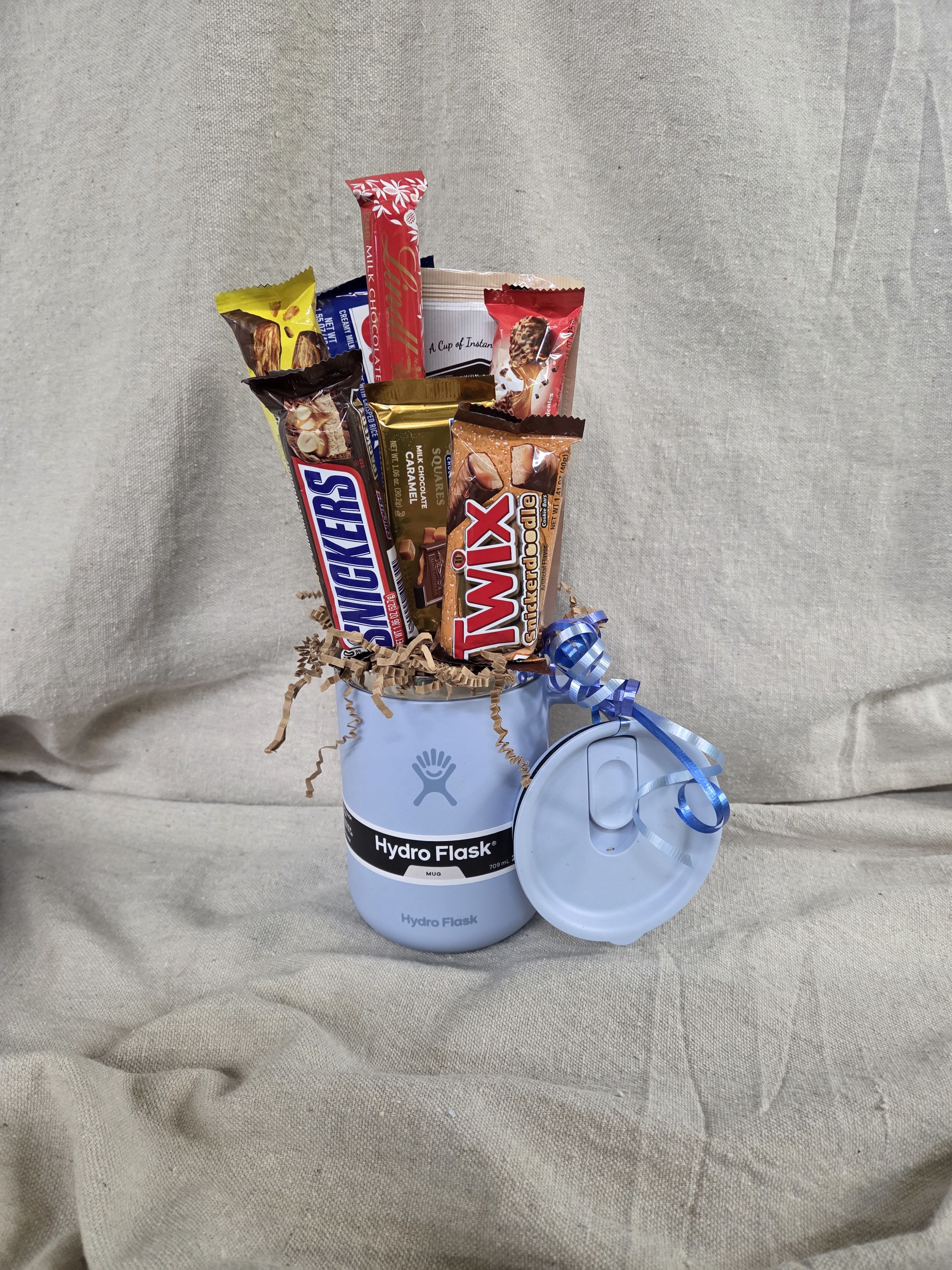 Light blue Hydro Flask mug filled with assorted candy bars and curly ribbon, with matching lid beside it.