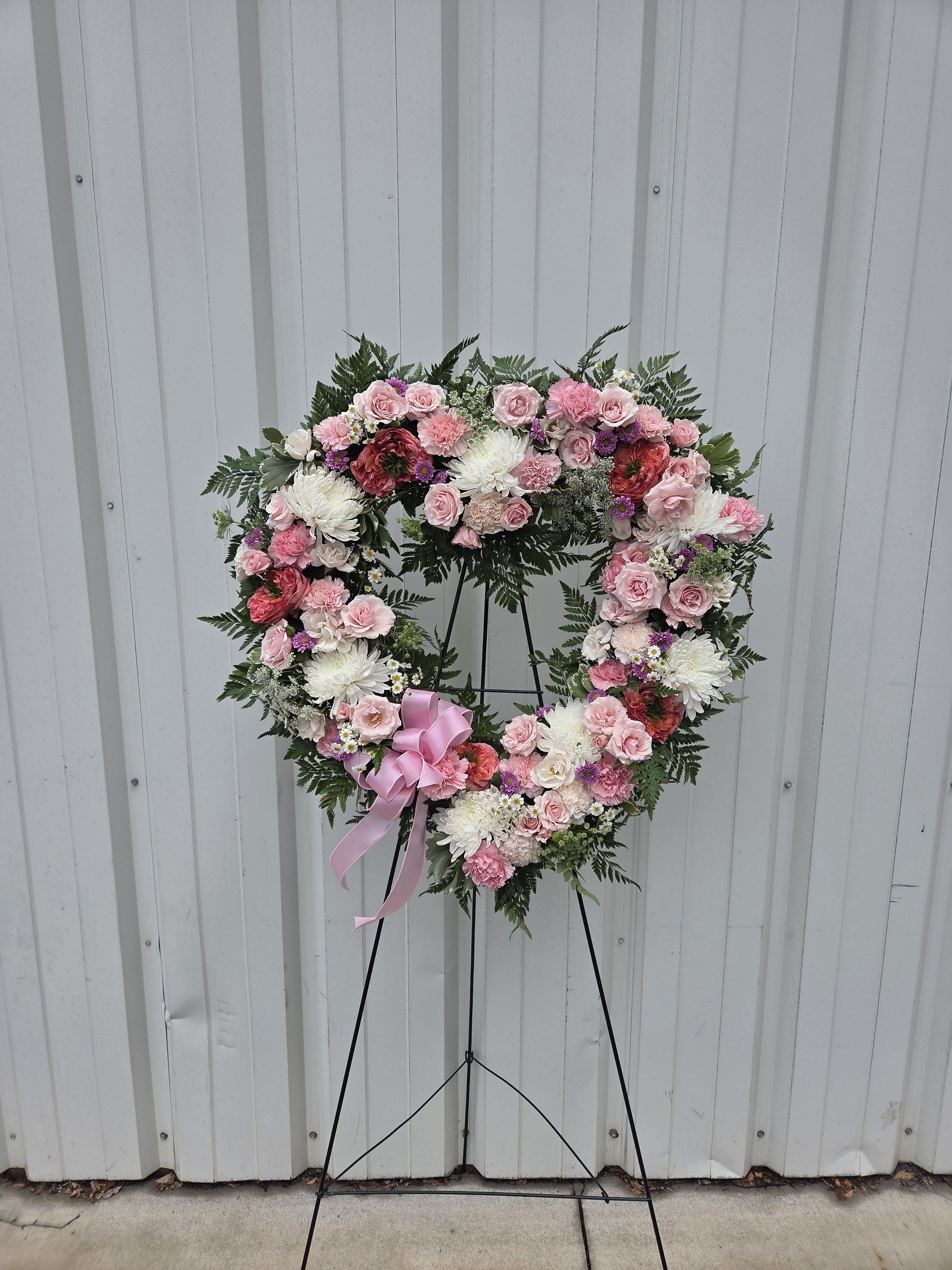 Heart-shaped standing wreath of pink and white flowers with a pink bow on an easel.