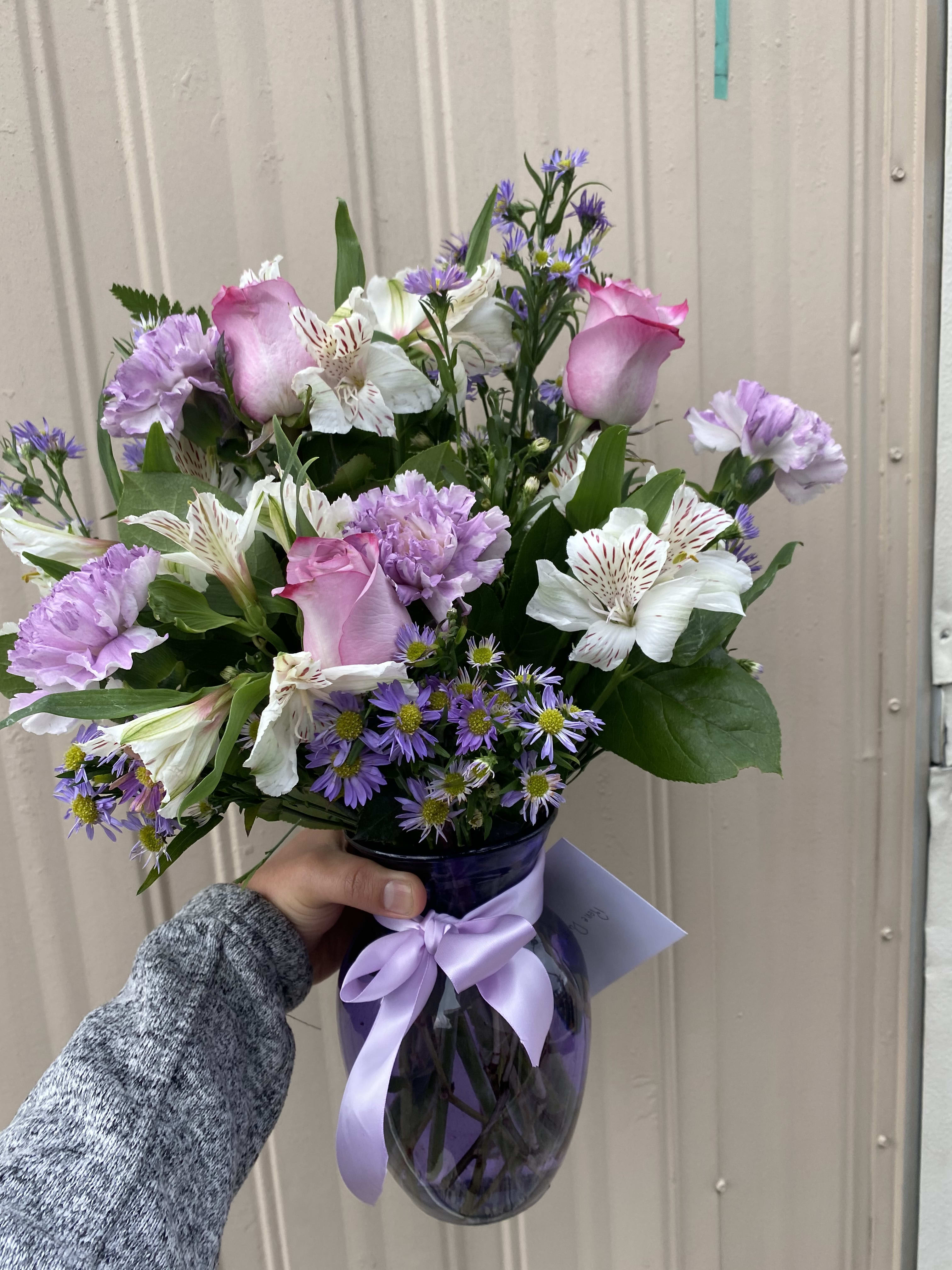 Pink roses and purple flowers arranged in a glass vase with a lavender ribbon.