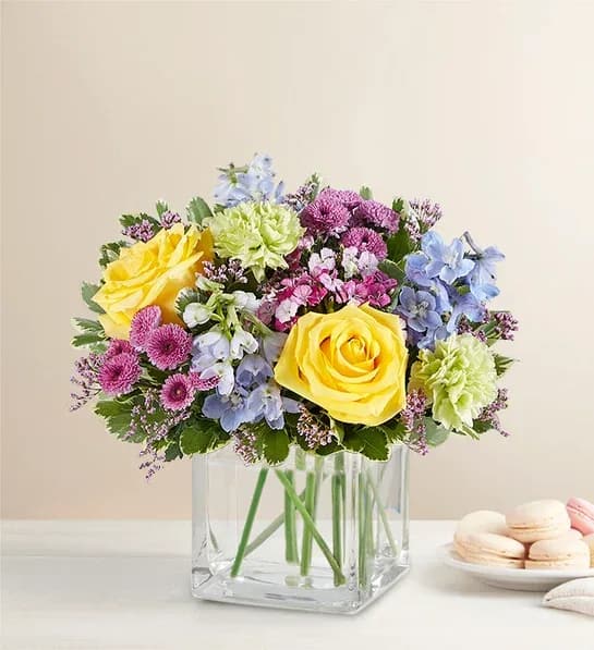 Bouquet of yellow roses, blue flowers, and pink blooms in a clear square vase