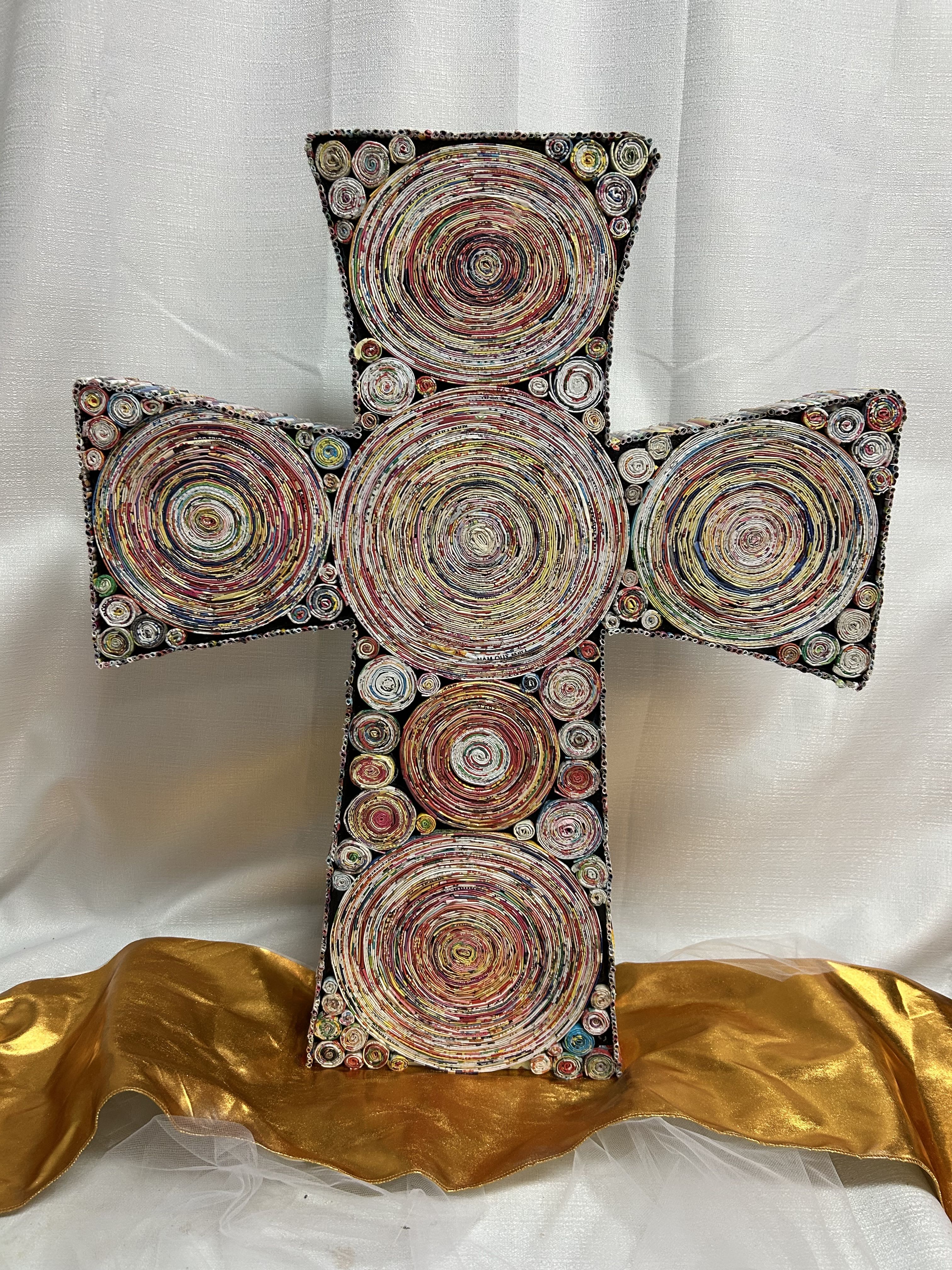 Multicolor decorative cross made of coiled circular patterns, set against white fabric with gold cloth at the base.