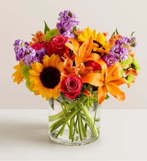 Mixed bouquet of sunflowers, roses, lilies, and purple flowers in a glass vase