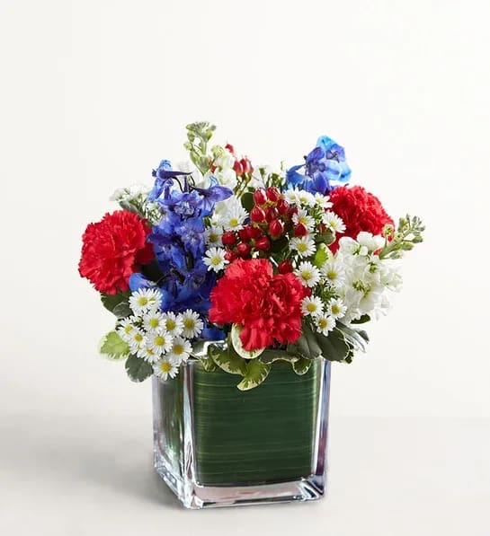 Healing Tears Red White &amp; Blue - It’s beautiful to honor one who held their country dear with a patriotic homage. Our arrangement of red roses, white stock, and blue delphinium, expertly gathered in a clear glass cube lined with Ti leaf ribbon, makes for a respectful and exquisite gesture of comfort and healing.