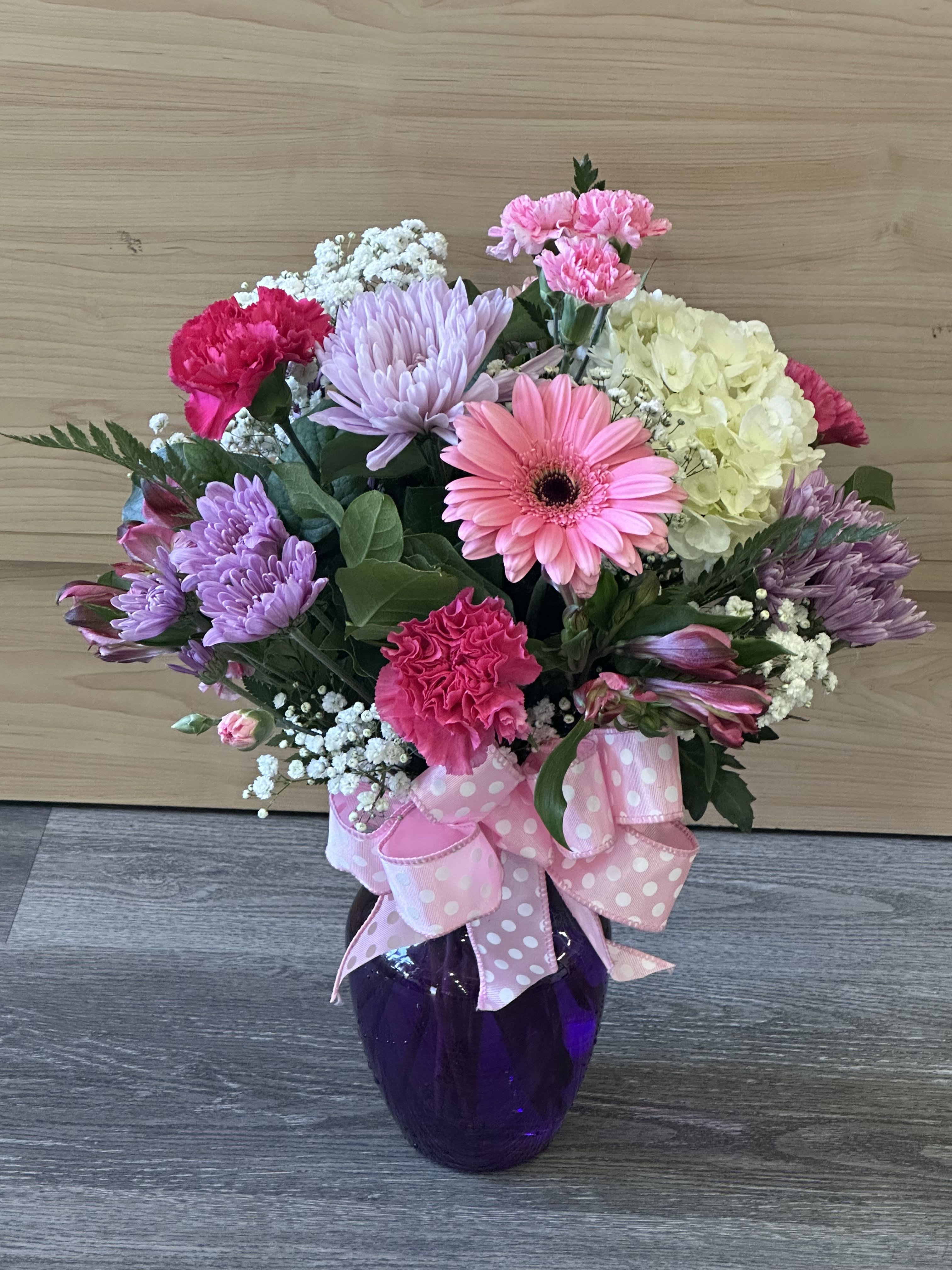 Mother’s Day bouquet - A lively, full-bodied Mother's Day bouquet that reads joyful and feminine at first glance-bright pink carnations and gerbera daisies mingle with lavender chrysanthemums, pale yellow hydrangea, creamy alstroemeria, and delicate white filler blooms to create a rounded, textured arrangement perfect for Mother's Day, birthdays, or a cheerful thank-you. The mix of ruffled carnations and large daisy-like blooms gives the bouquet an inviting, layered look that will make the person you're celebrating feel seen and adored.  Arranged densely in a deep purple glass vase and tied with a wide pink polka-dot ribbon, this floral gift arrives ready to display-no extra styling needed. Colors include pink, purple, white, and soft yellow; flowers include carnations, gerbera daisies, chrysanthemums, hydrangea, alstroemeria, and assorted filler blooms, giving it both visual variety and a soft spring scent. Ideal for moms, grandmothers, or any special woman who loves playful, feminine florals; order timed delivery to ensure peak freshness and immediate enjoyment.