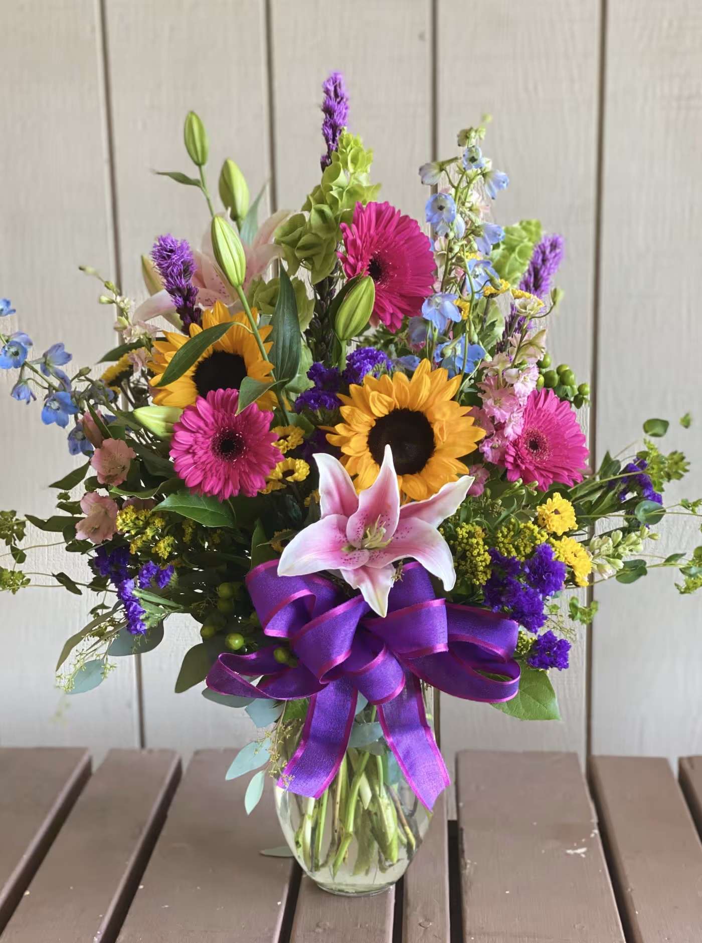 Summer Delight - Bright sunflowers and vivid pink gerbera daisies give this Summer Delight arrangement an instant burst of joy perfect for a birthday bouquet, a congratulations gift, or a cheerful just-because surprise. A prominent pink lily at the front draws the eye and adds a soft, fragrant focal point while purple spiky blooms and blue-and-yellow accents create lively contrast that keeps the composition feeling fresh and celebratory. The overall effect is energetic and upbeat-the kind of floral gift that makes the person you're thinking of feel genuinely seen and celebrated.  This tall mixed bouquet is gathered in a clear glass vase and tied with a large purple satin ribbon for a polished, festive finish. The combination of sunflowers, gerbera daisies, lilies, snapdragons and mixed filler flowers produces varied textures and layers-from the bold, sunny faces of the sunflowers to the delicate spikes and soft lily petals-so it reads beautifully from every angle. Ideal for brightening a table, office desk, or entryway; water daily and trim stems every few days to extend vase life.