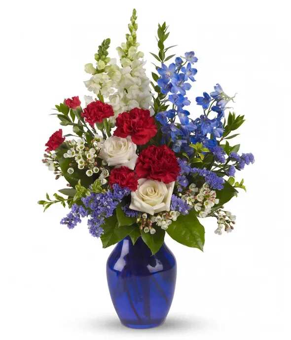 Freedom Remembrance - A striking upright bouquet built for remembrance and celebration, this patriotic arrangement is a perfect choice for Memorial Day, Veterans Day, or any occasion where you want to honor someone special. The freedom-themed centerpiece mixes red carnations and white roses with tall white snapdragon spikes and vivid blue delphinium-like blooms so the person you're thinking of will feel respected and remembered.  The design pairs glossy green foliage and small purple filler blossoms to add depth and a soft contrast around the central red, white and blue cluster. Delivered in a cobalt blue glass vase, the composition reads formal and celebratory-tall vertical lines from the snapdragons give it presence on a mantel or ceremony table while the roses and carnations provide lasting color.  Make this arrangement for a veteran, a grieving family, or to mark a patriotic holiday; it conveys honor, gratitude, and thoughtful remembrance. The vase helps keep stems neatly arranged and hydrated-simply change the water every few days and recut stems at an angle to extend freshness.