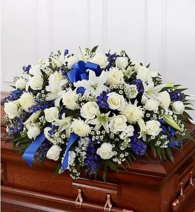 Blue &amp; white casket spray - A serene, formal casket spray crafted for a funeral or memorial service, this blue and white arrangement communicates respect and sympathy with quiet dignity. The standing spray and the low casket spray options both serve as a comforting tribute for a loved one, making it clear this is intended for memorial services and funeral services.  Made from white lilies, roses, daisies, carnations, snapdragons and chrysanthemums with spiky white accent blooms and broad patterned greenery, the arrangement pairs crisp whites with clusters of blue and purple accents for gentle contrast. Presented either tall on a floral easel or as a full, low spray to lay atop a polished wooden casket, the composition is symmetrical and layered, with blue ribbon loops adding a ceremonial finish.  Perfect for honoring a parent, spouse, friend, or colleague, this spray strikes a solemn, traditional tone that helps the person you're remembering feel celebrated and respected. Available in the standing easel format or the casket-top style-please specify placement and any personalization needs when ordering so we can ensure the appropriate size and ribbon detail for the service.