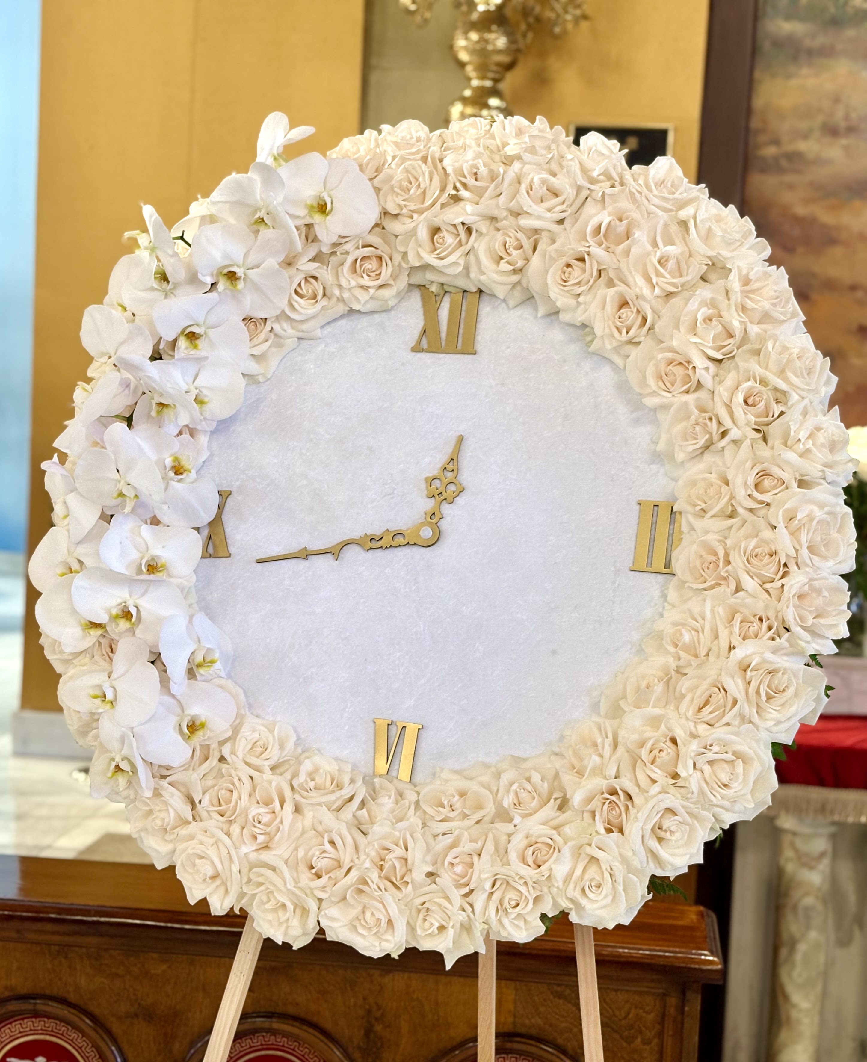 " The Clock " By VLM Flowers - A refined circular wreath of cream roses and white orchids arranged around a functioning clock face creates a dignified memorial flower arrangement for sympathy and remembrance. This standing tribute is crafted as a formal sympathy arrangement suitable for a funeral service, graveside memorial, or a tribute display to honor someone who has passed-an elegant way to show the person you're thinking of that their memory is cherished.  Dense cream roses form the broad, soft ring while clusters of white orchids accent one side, lending a gentle asymmetry and delicate texture against the smooth clock dial. The palette of white, cream, and soft pink keeps the presentation restrained and respectful; mounted on an easel with the clock at center, the piece reads as both floral art and a meaningful keepsake. The substantial size and symmetrical composition ensure it displays well at a service or reception.  This arrangement sends a clear message of remembrance and calm-an honoring gesture that helps loved ones feel seen and comforted. Care is minimal for the life of the display: keep away from direct heat and replace water as needed while on-site; please allow appropriate lead time when ordering to ensure timely delivery to the venue.