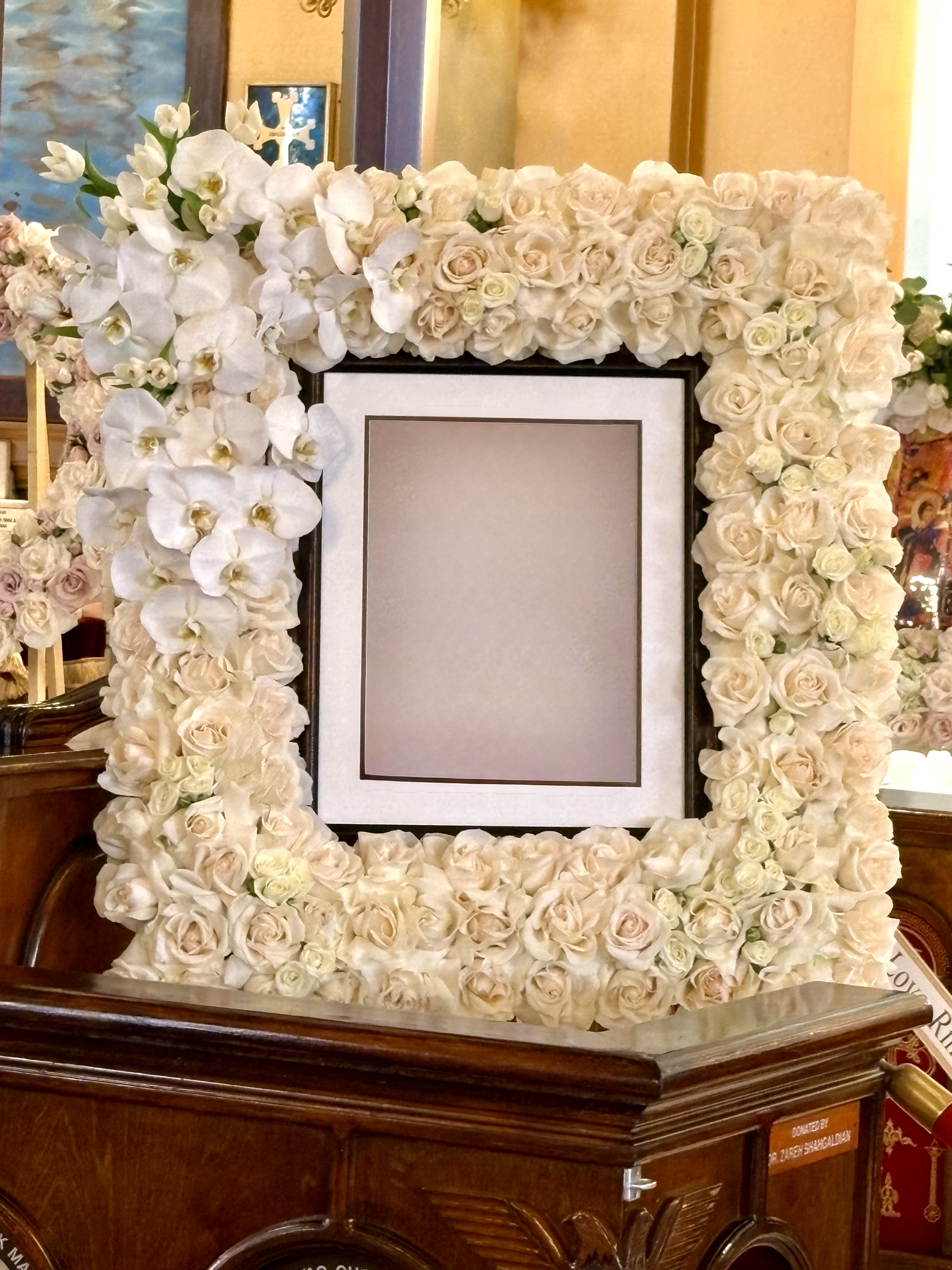 Forever in Bloom By VLM Flowers - A soft ivory floral frame built from white Phalaenopsis orchids and cream-to-blush roses creates a serene, formal focal point for a memorial display. This picture frame arrangement frames a central photo opening with dense, symmetrical blooms so the person you're honoring .  The design features clusters of white phalaenopsis orchids along the left with cream-to-blush roses filling the top, right and bottom edges, producing a polished, cohesive look in white, pink and green tones. The arrangement is compact and neatly edged to sit around a standard framed photo; textures range from the orchids' glossy, sculptural petals to the soft, layered rose blossoms, giving visual depth without overwhelming the image it surrounds.  