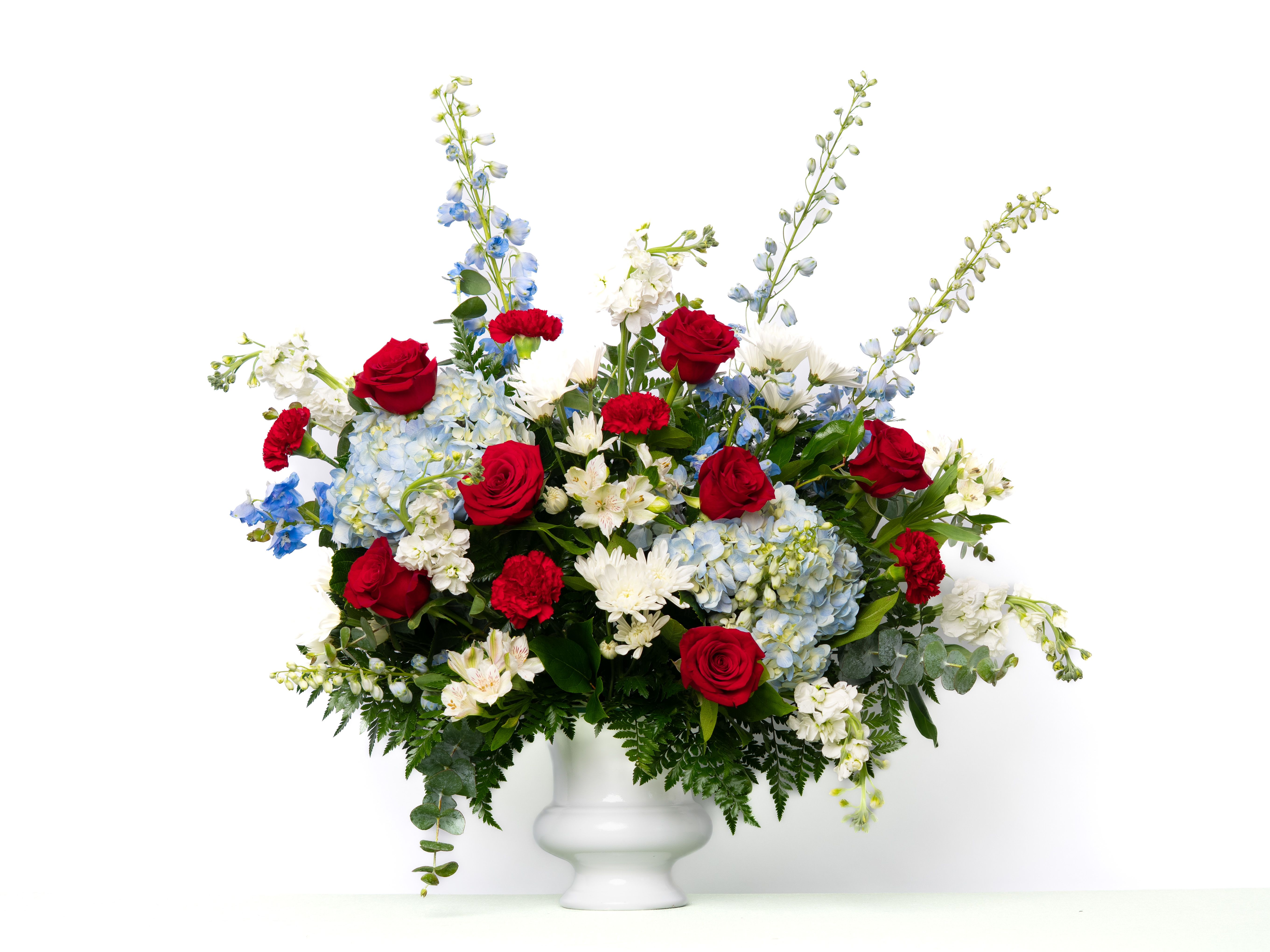 Our Honor Collection: Tribute Urn in Red, White, and Blue Blooms - A meaningful tribute honoring a life of service, love, and legacy. Designed in a patriotic palette of red, white, and blue, this arrangement is a respectful and dignified way to celebrate and remember.  Each arrangement is thoughtfully designed using the freshest blooms available. Flower varieties may vary based on availability while maintaining the overall style, color palette, and value. Urn styles and colors may change based on weekly availability and will be substituted to maintain the overall value.  Approximate Dimensions of Floral Tribute: 30" tall x 33" wide  Color &amp; Customization: Need a custom color scheme? Contact us for more information. Our most popular palettes include whites, whites and greens, whites and blues, pinks and whites, pastel shades, and colorful designs.  Delivery: All sympathy pieces are hand-delivered by the Schultz Floral team.