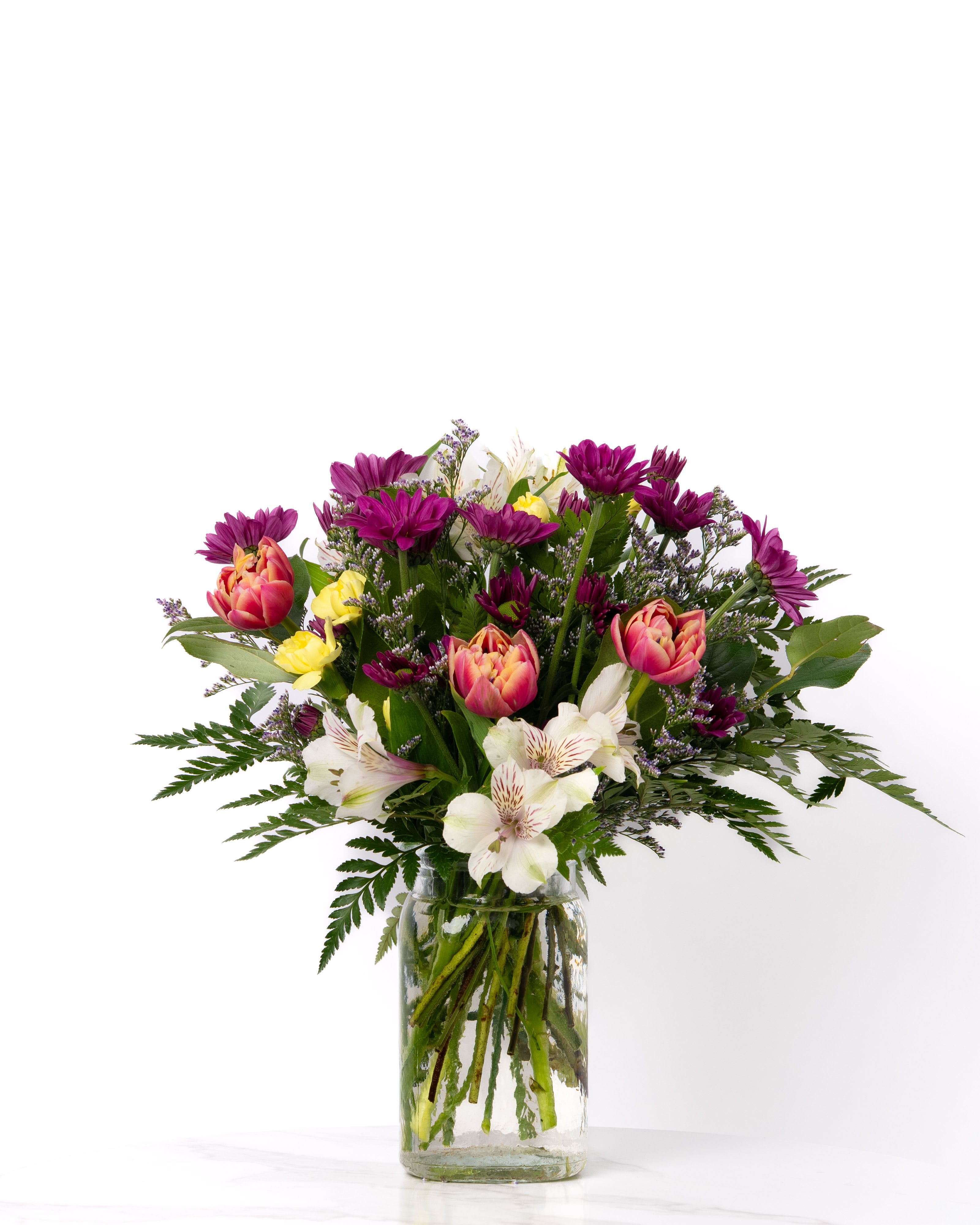 Mixed bouquet of purple daisies, pink tulips, and white blooms in a clear glass vase