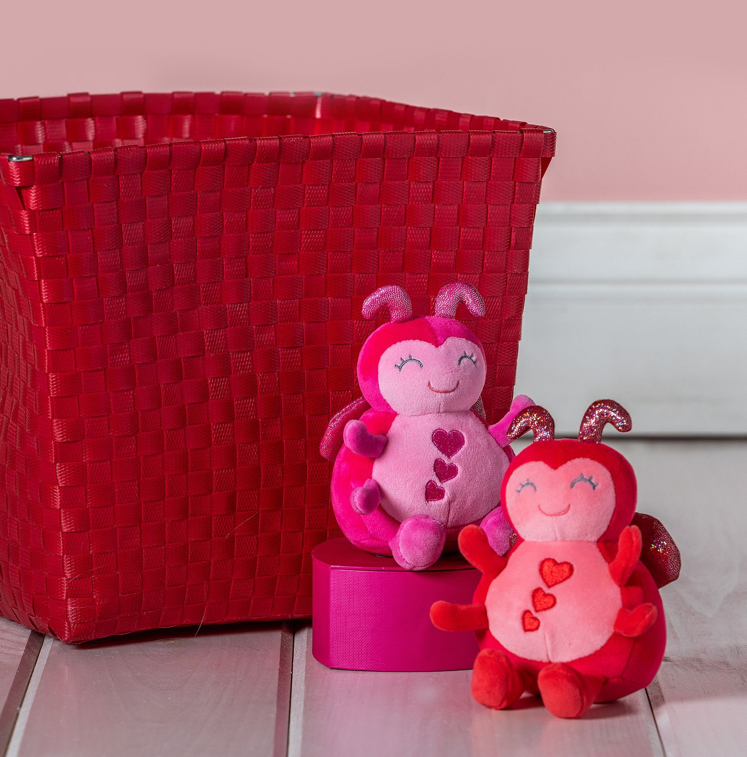 Two pink and red heart-themed plush ladybug toys beside a red woven basket