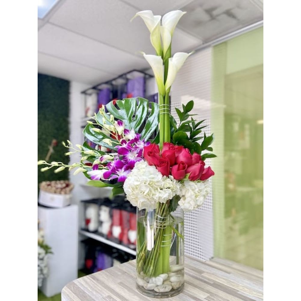 Tall modern arrangement with calla lilies, orchids, roses and hydrangeas in a glass vase with a monstera leaf