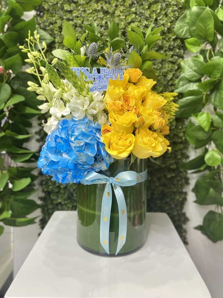 Tall glass vase with yellow roses, blue hydrangea, white orchids and an 'It’s a Boy' pick.