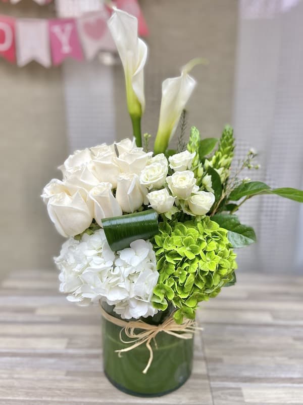 Tall white and green arrangement with calla lilies, roses, and hydrangeas in a glass cylinder vase
