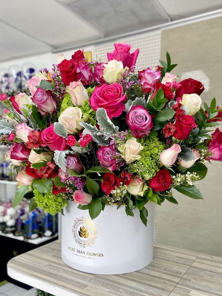 Large arrangement of red, pink, and ivory roses with green accent blooms in a white hatbox container