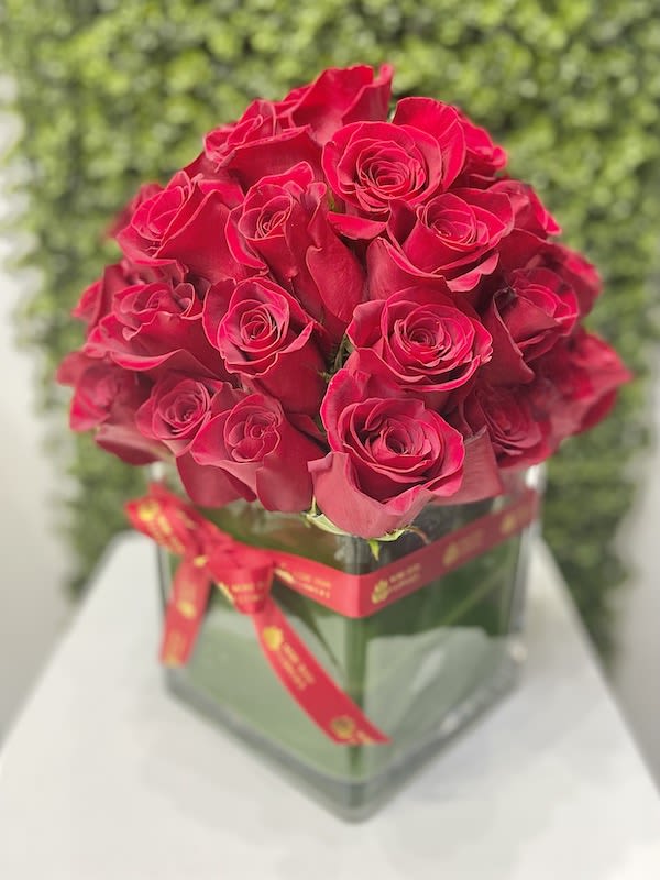 Dense bouquet of red roses arranged in a square glass vase wrapped with a red ribbon.