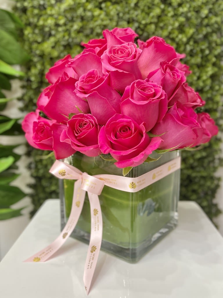 Compact arrangement of bright pink roses in a square glass vase with a pale pink ribbon bow.