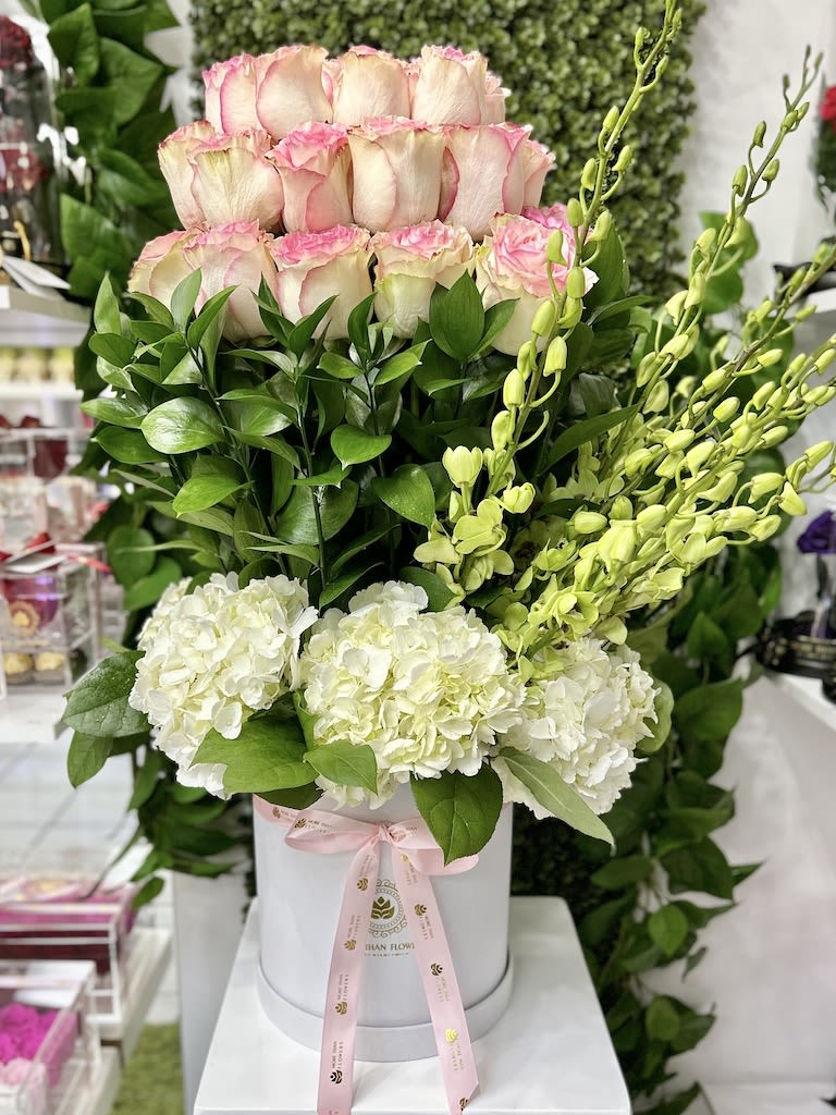 Tall arrangement of pink-tipped roses, white hydrangeas, and green orchids in a white hatbox with pink ribbon