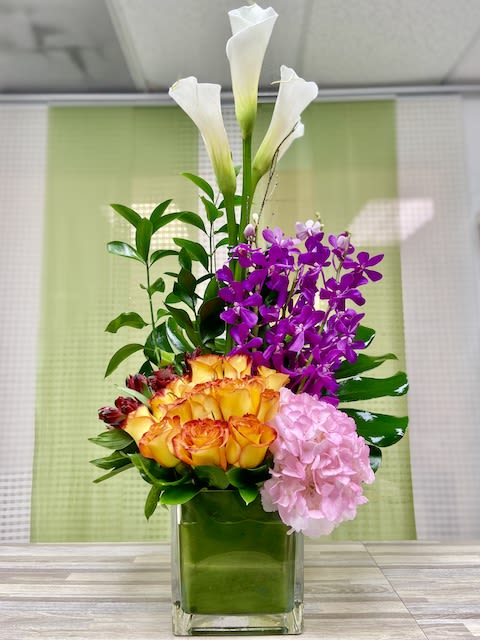 Tall modern arrangement with yellow roses, purple orchids, white calla lilies, and pink hydrangea in a glass cube vase