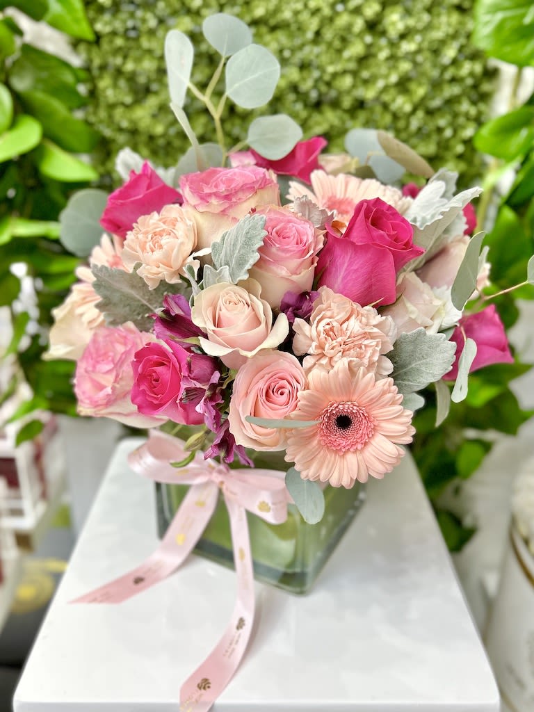 Pink and peach roses, carnations, and gerbera daisies in a glass cube vase with a pink ribbon bow