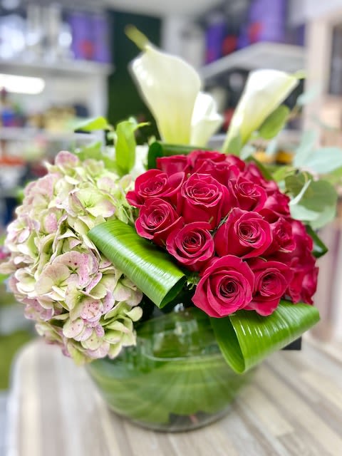 Low glass vase arrangement with hot pink roses, pale hydrangea, white calla lilies, and folded green leaves.