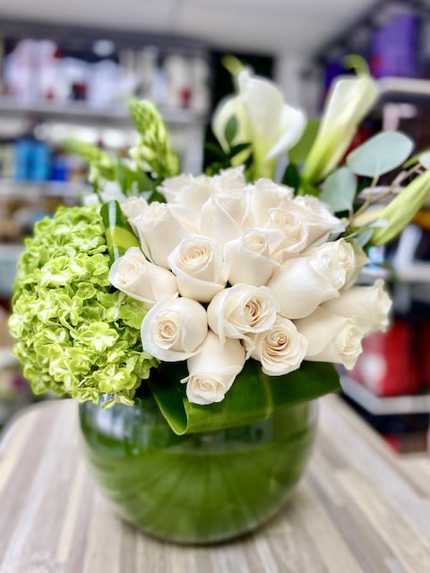 Round glass vase filled with white roses, white calla lilies, and green hydrangeas.