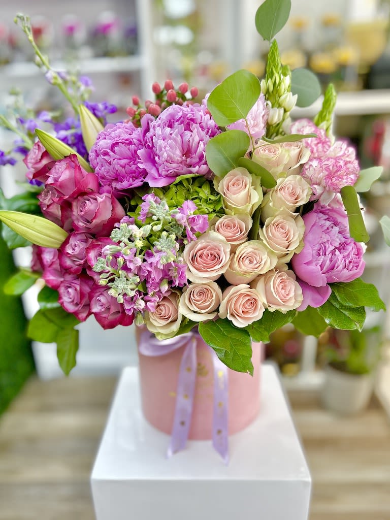 Pink and blush roses, peonies, and lilies arranged in a round pink hatbox with ribbon