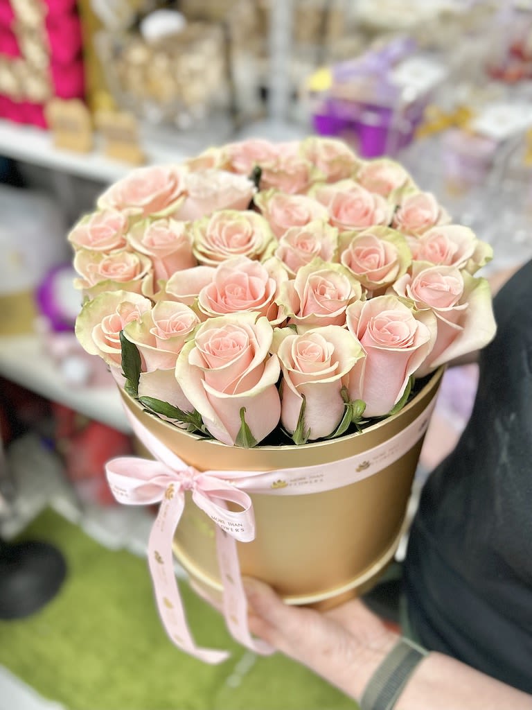 Round gold hat box filled with tightly packed pale pink roses and tied with a pink ribbon.