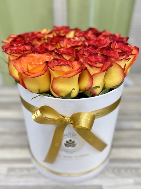 Round box of yellow and orange-red roses with a gold ribbon bow on a white container