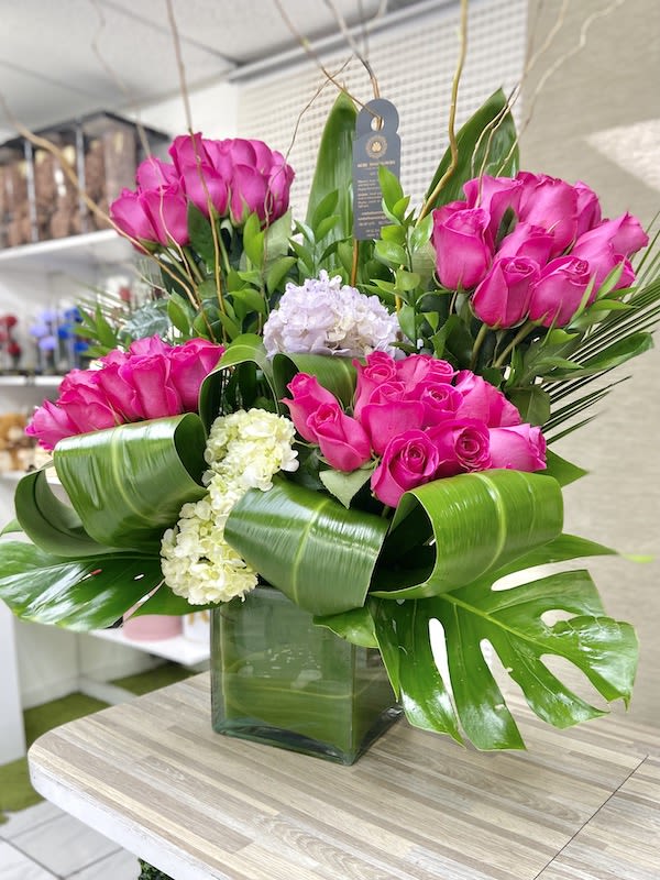 Tall arrangement of hot pink roses with white hydrangeas and large green tropical leaves in a glass cube vase