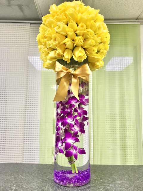 Tall glass vase with submerged purple orchids and a dome of yellow roses tied with a satin bow.