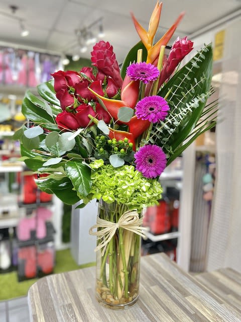 Tall tropical arrangement with red roses, orange blooms, green hydrangea and purple daisies in a glass vase