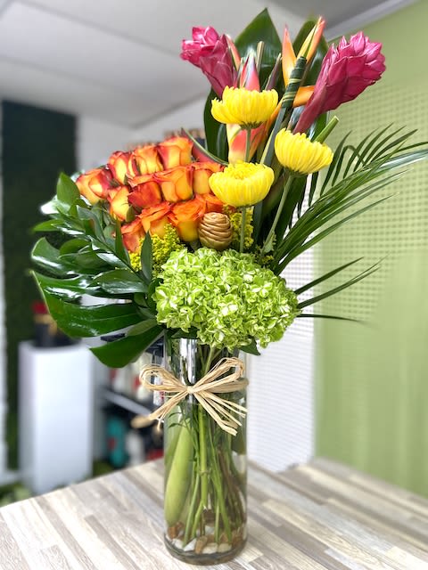 Tall tropical arrangement with orange roses, green hydrangea and yellow blooms in a glass vase with raffia bow