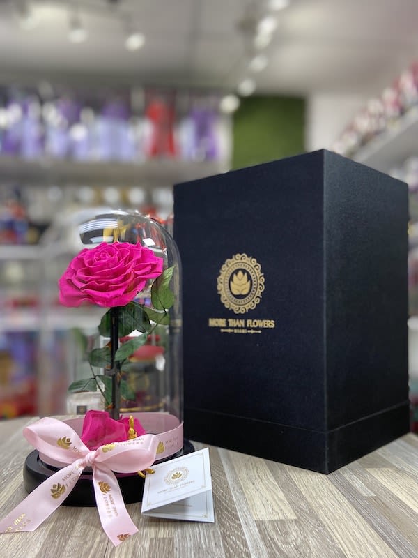 Single bright pink rose under a glass dome on a black base with a tall black gift box beside it.