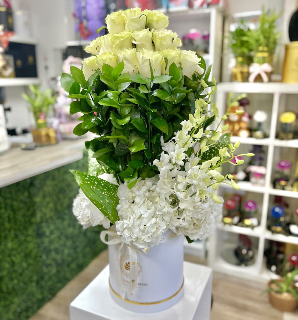 Tall arrangement of pale roses, white blooms, and dense greenery in a white hatbox container