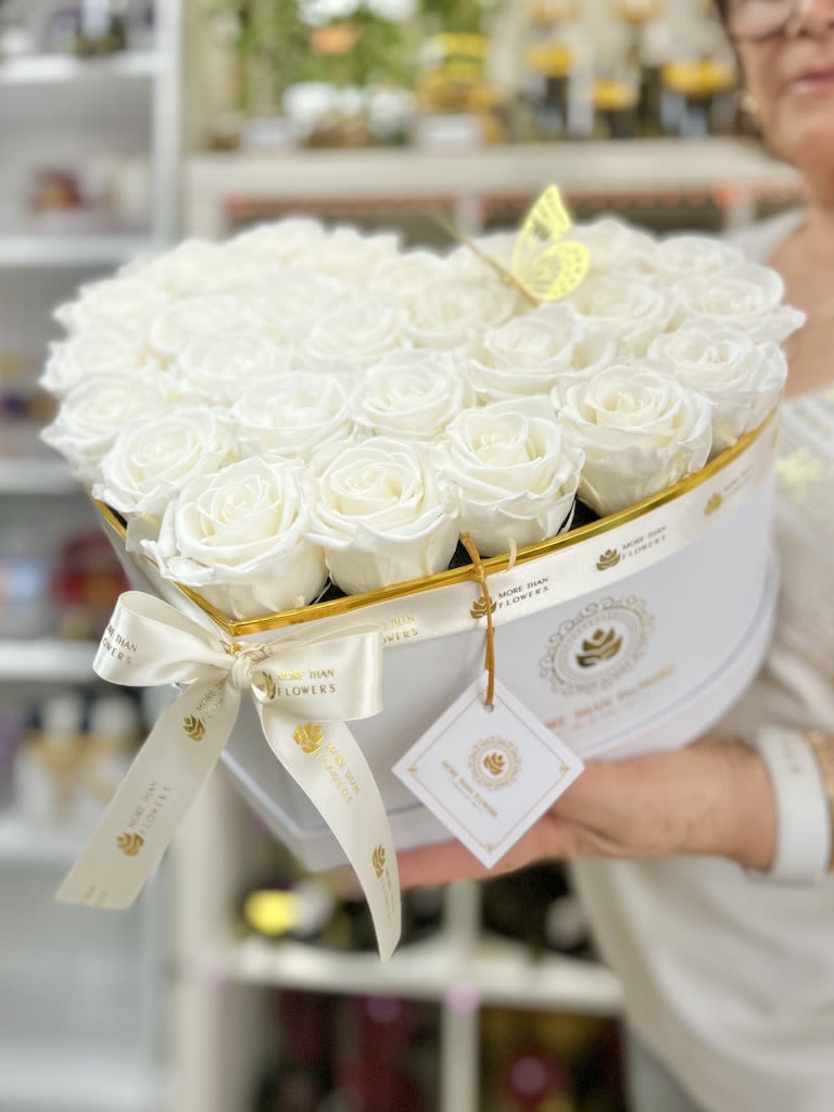 Heart-shaped white box filled with white roses, tied with a white ribbon and tag.