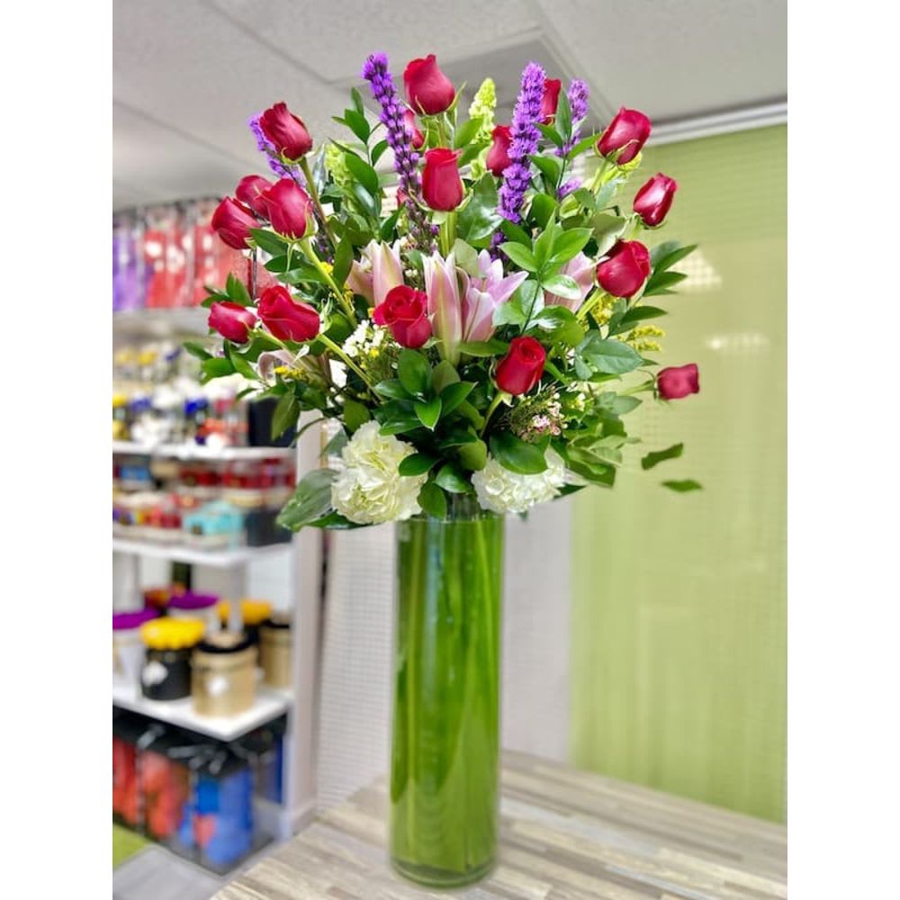 Tall arrangement of red roses, pink lilies, white carnations and purple flowers in a green glass cylinder vase